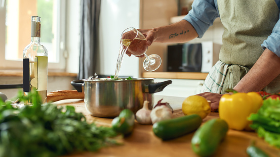 Cropped shot of man, chef cook pouring a glass of white wine into the pan with chopped vegetables while preparing a meal in the kitchen. Cooking at home, Italian cuisine. Selective focus. Web Banner