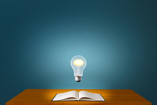 Glowing light bulb in mid-air over the desk with open blank book against blue concrete wall with copy space.