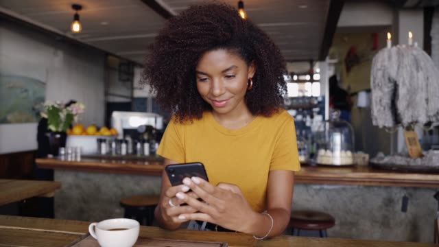 African American smiling female typing to boyfriend on smartphone sitting in trendy cafe next to hot cup of coffee