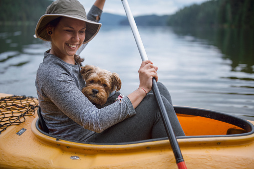 Cheerful smiling woman is kayaking with her cute terrier dog at the lake with a beautiful view at the background