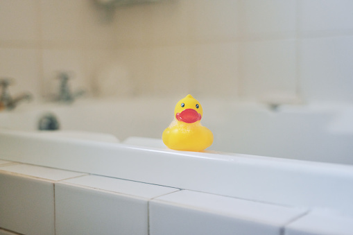 A yellow duck on the baby's head. the baby is sitting in the bathroom with foam. bathroom games