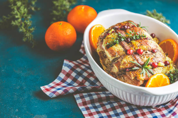 Roasted pork in white dish, christmas baked ham with cranberries, tangerines, thyme, rosemary, garlic on dark blue table surface stock photo