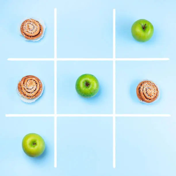 Green apples vs cinnamon buns in tic tac toe or noughts and crosses game, healthy vs unhealthy food concept, top view, square format