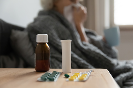 Close up pills bottles and packaging on bedside table in mature woman bedroom, unhealthy sick middle aged female suffering from flu, cold or seasonal virus, allergy, recovery and treatment concept
