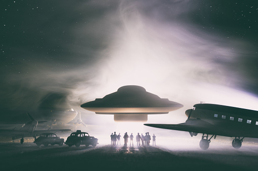 An old-style UFO hovers over an airfield, flanked by a Spitfire and a couple of DSC-3 Dakotas. A group of people look on in wonder. Miniature photography.