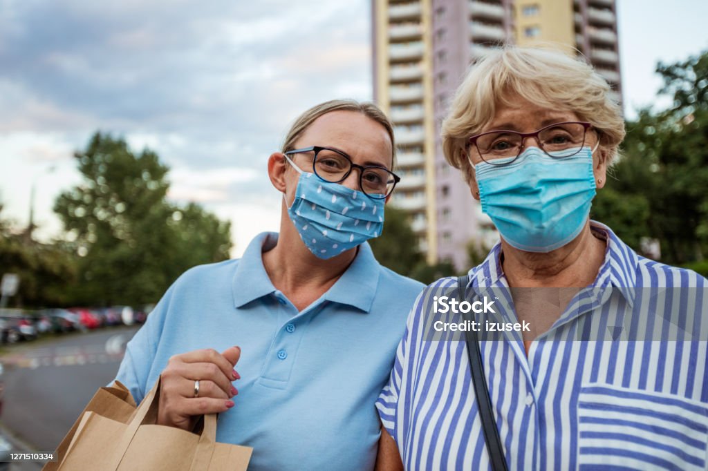 Home caregiver helping senior woman with shopping Outdoor portrait of senior woman and social worker. They are wearing protective face masks, standing in front of apartment in the city. 70-79 Years Stock Photo
