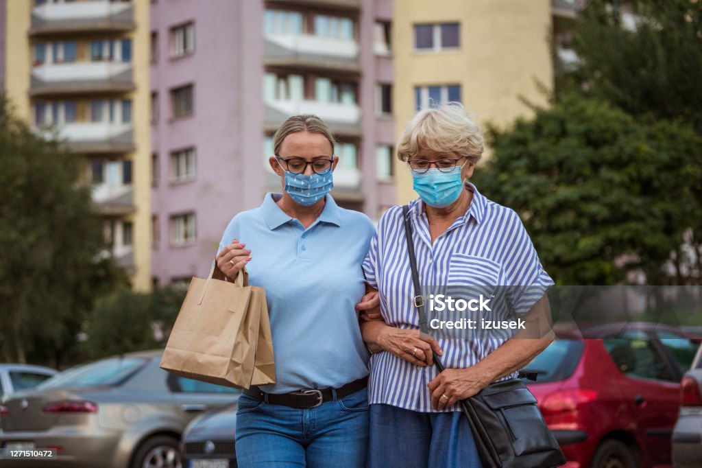 Home caregiver helping senior woman with walking outdoors Outdoor shot of senior woman and female social worker. They are wearing protective face masks, walking in front of apartment in the city. 70-79 Years Stock Photo