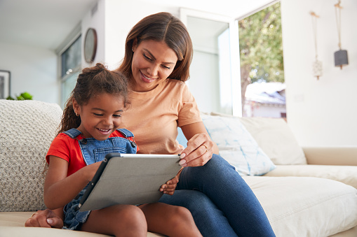 Hispanic Mother Helping Daughter To Home School And Do Homework  With Digital Tablet