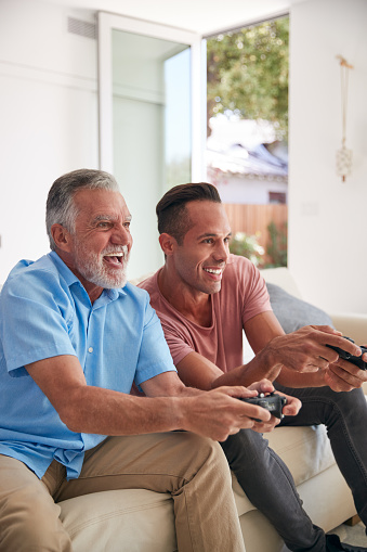 Hispanic Father With Adult Son Sitting On Sofa At Home Playing Video Game Together