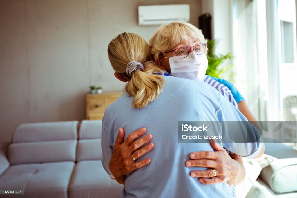 Home caregiver supporting senior woman during coronavirus pandemic District nurse embracing worried with senior woman during home visit. They are standing in living room. Embracing Stock Photo