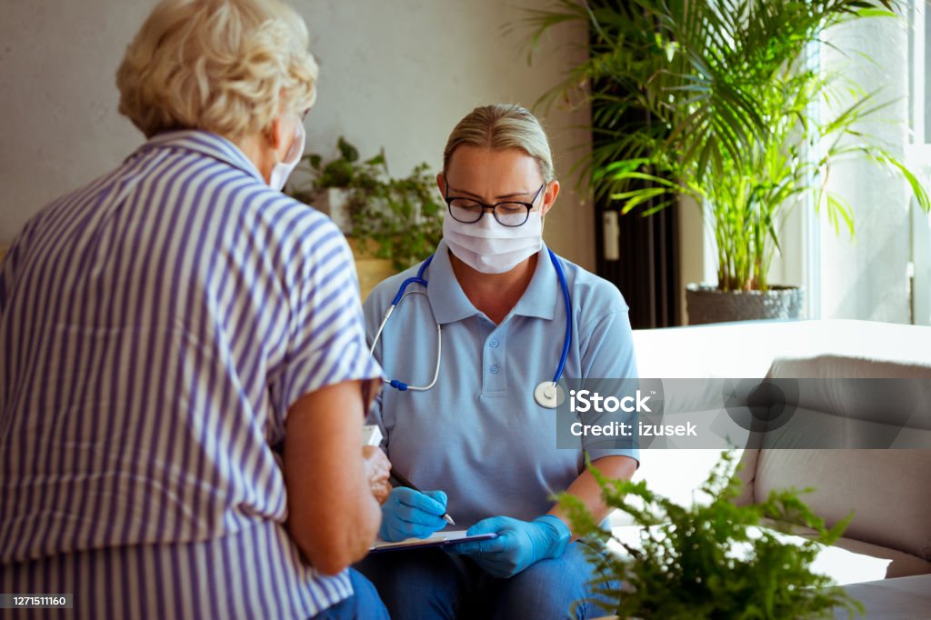 Home caregiver visiting senior woman during coronavirus pandemic District nurse wearing protective face mask and gloves talking with senior woman during home visit. They are sitting in living room. Home Caregiver Stock Photo