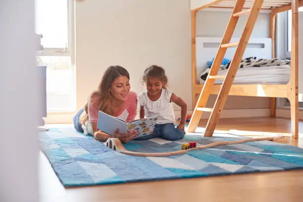 Photo of Hispanic Mother And Daughter Reading Book And Playing With Toy Train Set In Bedroom Together