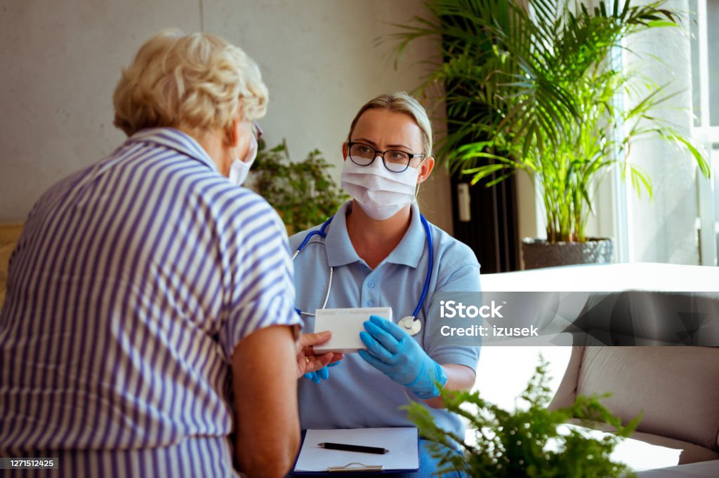 Nurse talking with senior woman during home visit District nurse wearing protective face mask and gloves talking with senior woman during home visit, holding pills. They are sitting in living room. Diabetes Stock Photo