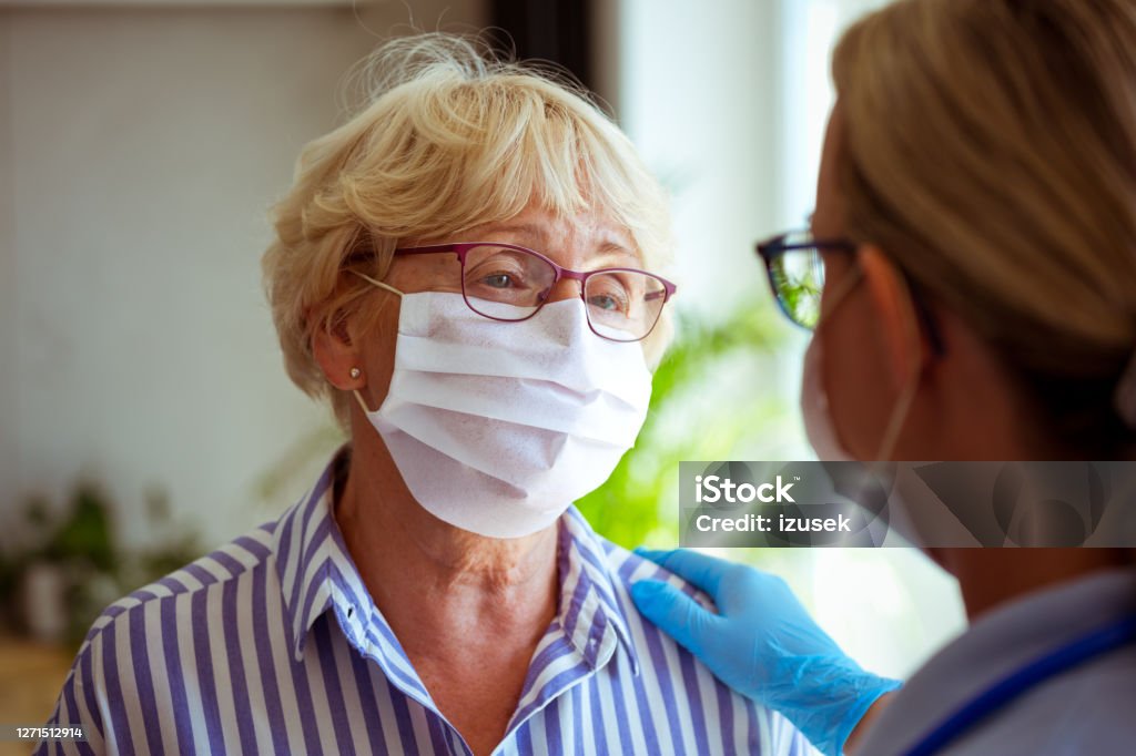 Worried senior woman talking with home caregiver Sad senior woman wearing protective face mask talking with district nurse during coronavirus pandemic. They are standing in living room. Close up of faces. Doctor Stock Photo