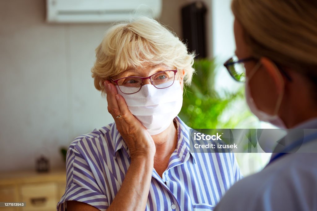 Worried senior woman talking with home caregiver Sad senior woman wearing protective face mask talking with district nurse during coronavirus pandemic. They are standing in living room. Close up of faces. Senior Adult Stock Photo