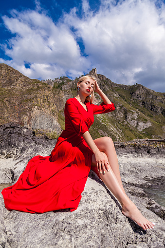 A blonde woman in a red long dress sits on the stone bank of the river against the backdrop of mountains with closed eyes and make-up on her face and runs her hand along her outstretched long legs.