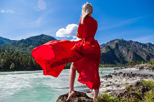 Blonde woman in long red dress fluttering in wind stands barefoot on rocky shore with one leg outstretched against background of mountains, blue clouds and birch-tree river in Altai. View from back