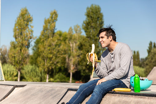boy with roller skates eating a banana sitting in a public skate park. he has a protective helmet and a bottle of water on the side