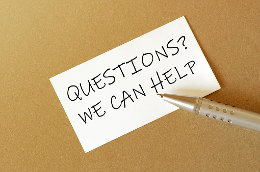 pen, white paper with text Questions We Can Help on the brown background