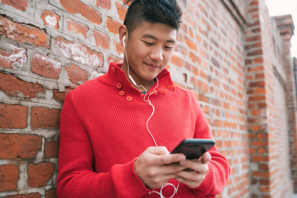 Portrait of young Asian man using his mobile phone with earphones outdoors against brick wall. Communication concept.