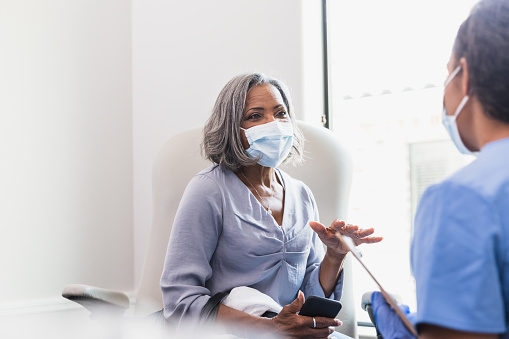 An attentive female triage nurse listens as a senior female patient describes her symptoms. The patient and nurse are wearing protective face masks.
