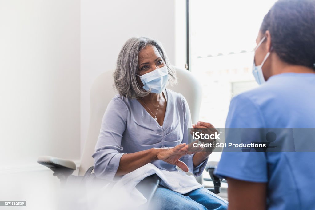 Senior woman talks with female healthcare professional A senior woman, wearing a protective face mask, talks with a female nurse during a medical appointment. Doctor Stock Photo