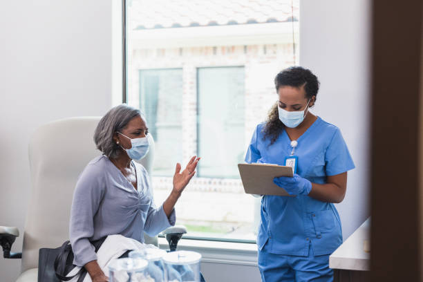 Senior woman gestures while talking with nurse A female nurse takes notes as a senior female patient discusses health concerns. The patient and nurse are wearing protective face masks. triage stock pictures, royalty-free photos & images