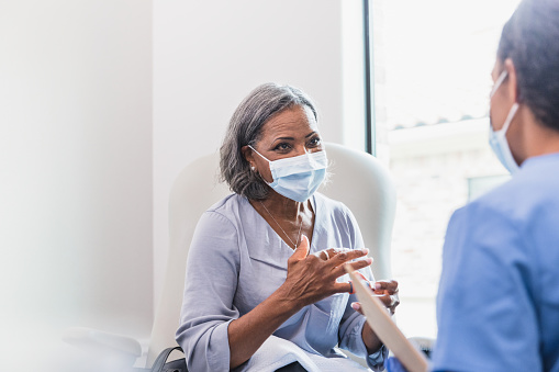 A senior female patient gestures while describing her symptoms to a female healthcare provider. The patient and doctor are wearing protective face coverings.