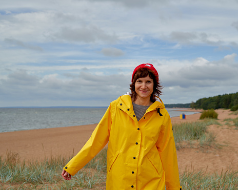 Waist portrait of mature woman in yellow raincoat and red hat walking along beach and looking at camera, smiling. Autumn walk in nature in bright clothes. Female balancing on the beach