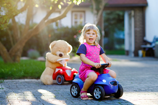 Little adorable toddler girl driving toy car and having fun with playing with plush toy bear, outdoors. Gorgeous happy healthy child enjoying warm summer day. Smiling stunning kid in gaden.