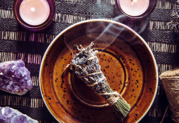 Homemade herbal lavender (lavendula) smudge stick smoldering on white plate with candles and amethyst crystal clusters for decoration. Spiritual home cleansing concept. Homemade herbal lavender (lavendula) smudge stick smoldering on white plate with candles and amethyst crystal clusters for decoration. Spiritual home cleansing concept. incense photos stock pictures, royalty-free photos & images