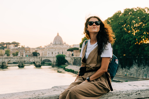 Portrait of a young adult beautiful woman sitting on a bridge in Rome, Italy. She's looking away at sunset.