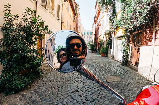 Young couple taking a picture on the rearview mirror of a red motor scooter in Rome, Italy. Trastevere district.