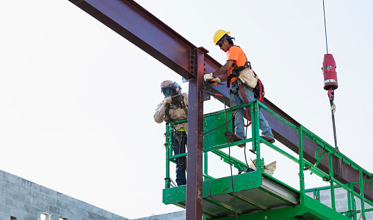A welder working on attaching a steel girder to a column at a construction site. The young man is standing on a scissor lift, face obscured by his welding mask. Another construction worker, a mid adult Hispanic man, is working next to him on the other side of the column.