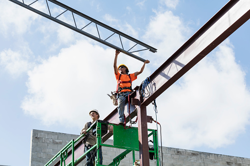 Two multi-ethnic construction workers on a scissor lift. They are steel workers installing a roof joist. The one wearing a yellow hardhat is an Hispanic mid adult man in his 30s. His coworker is a young man in his 20s.