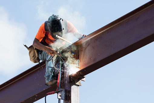 An Hispanic steel worker working high up on a girder. He is sitting on the girder, wearing a safety harness, welding to secure the girder to a column.