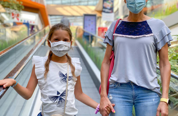 保護フェイスマスクを身に着けているショッピングモールで彼女の母親と手をつないで陽気な小さな女の子 - ストック写真 - escalator child shopping mall little girls ストックフォトと画像