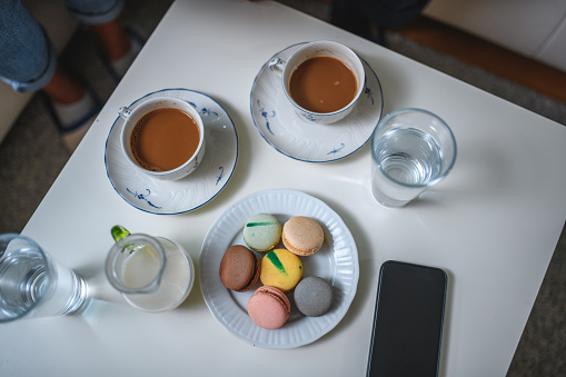 Unrecognisable people drinking coffee indoors in a living room together. They are eating macaroons. Directly above.