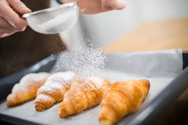 Chef sprinkling powdered sugar on croissants Close-up of chef's hand sprinkling powdered sugar on croissants with sieve in kitchen. sprinkling powdered sugar stock pictures, royalty-free photos & images