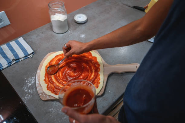 Hispanic man preparing pizza in domestic kitchen. He is putting tomato sauce on the dough.