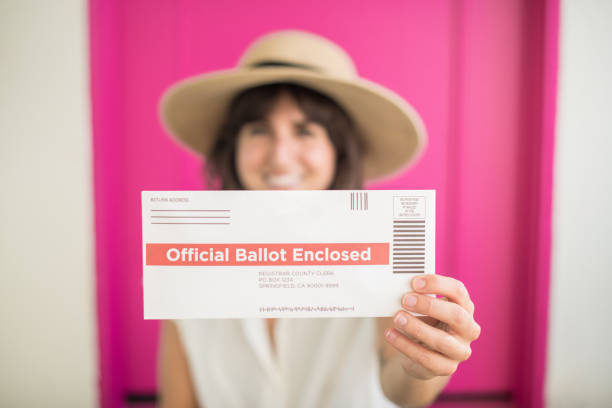 A young woman holds her completed absentee voter ballot ready to be mailed in for the 2020 presidential election while standing in front of her pink door.