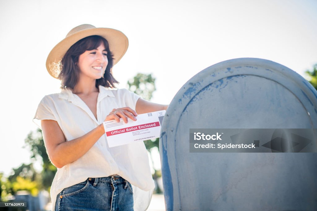 Woman Mailing Her Absentee Voter Ballot A young woman places her absentee voter ballot for the 2020 presidential election into a blue United States Postal mailbox. Mailbox Stock Photo