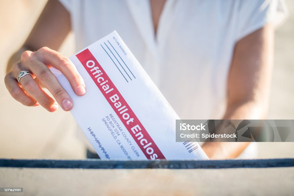 Woman Mailing Her Absentee Voter Ballot A young woman places her absentee voter ballot for the 2020 presidential election into a blue United States Postal mailbox. Voting Ballot Stock Photo
