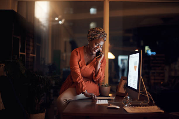 I just called to see if you can help me with something... Shot of a young businesswoman talking on a cellphone while using a computer in an office at night leanincollection stock pictures, royalty-free photos & images