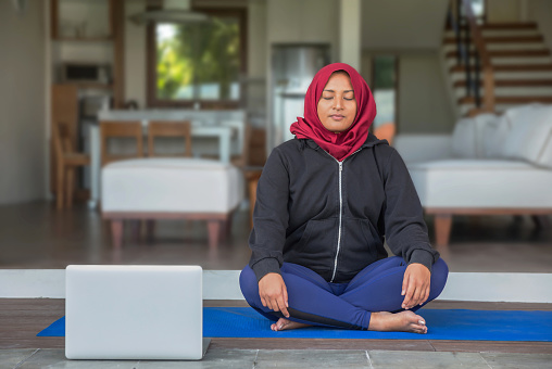 Portrait shot of a Malaysian muslim woman with hijab, sitting in a lotus position, closing her eyes while listening morning meditation guide from her laptop.