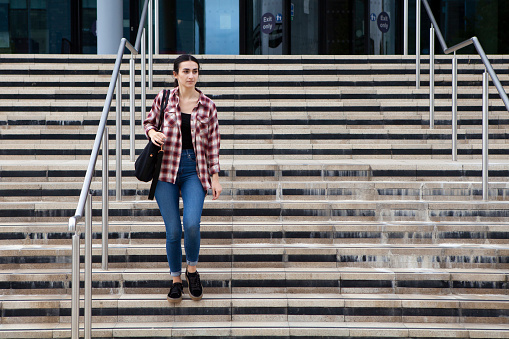 Female university student wearing a checked shirt and jeans, and carrying a shoulder bag, walking down steps from a university campus building
