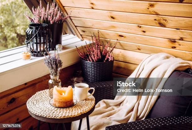Cute Autumn Home Decor Arrangement Tiny Wooden Cabin Balcony With Heather Flowers In Pot Lavender In Bottle Vase Candlelight Flame Soft Beige Plaid Waiting On Comfortable Garden Furniture Chair Stock Photo - Download Image Now