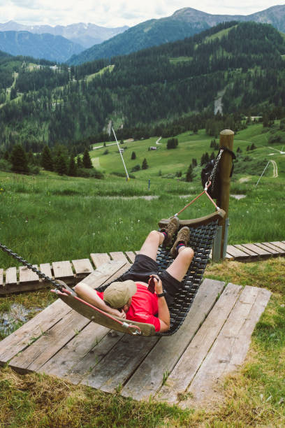 彼の下に素晴らしい景色を持つ山の木製のハンモックに横たわっている赤いシャツを着た男 - 緑の谷の美しい景色 - 休憩を取り、オーストリアのアルプスでのハイキング旅行で休んでいます� - footpath field nature contemplation ストックフォトと画像