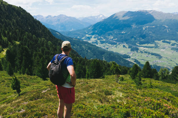 Rear view of fit mature man looking down to a beautiful valley while standing on the mountains in summer on his hiking vacation. Strong, brave, active, healthy lifestyle, feeling of adventure and freedom. stock photo