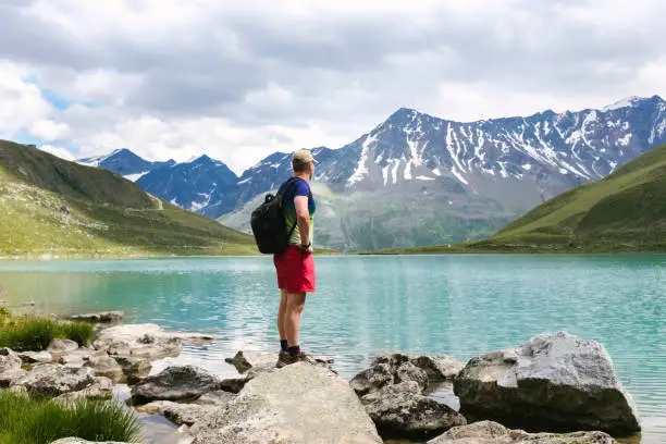 July 21, 2020 - Rifflsee, Austria: Man enjoying the view of an emerald lake in the mountains reflecting snow on the peaks - hiking summer vacations in Austrian Alps - feeling of adventure, freedom, getting away from it all, standing on the top of the world.